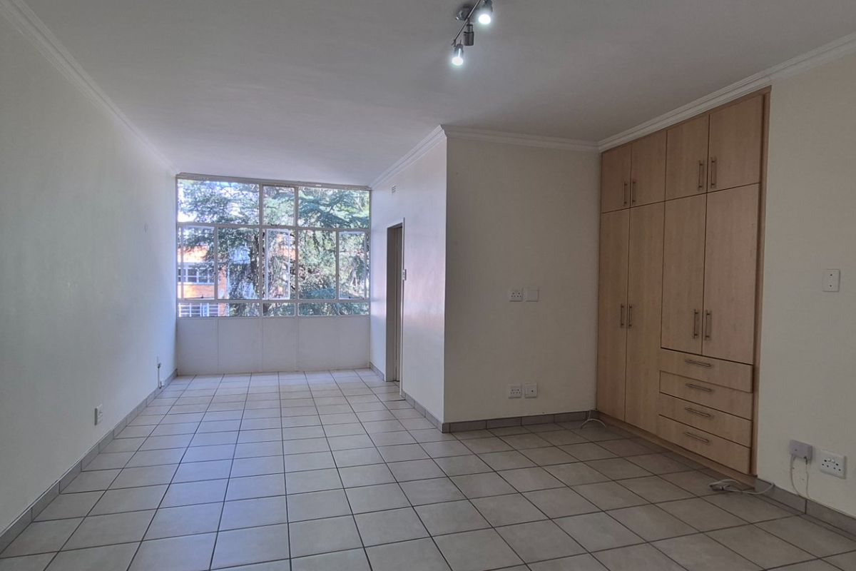 A bright, empty bedroom featuring neutral tiled flooring, built-in wooden cupboards, and a large window providing natural light.