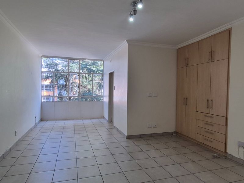 A bright, empty bedroom featuring neutral tiled flooring, built-in wooden cupboards, and a large window providing natural light.