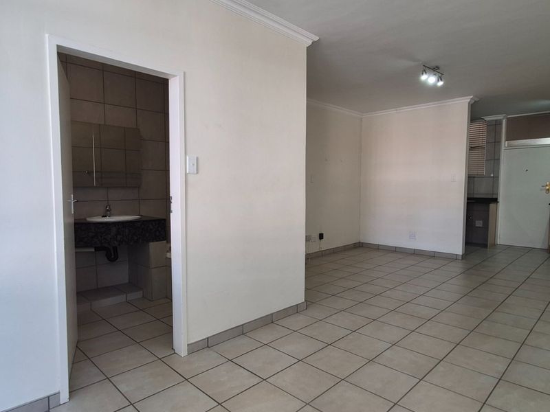 An open-plan living area featuring tiled floors, coving, and a view into a functional kitchen and bathroom space.