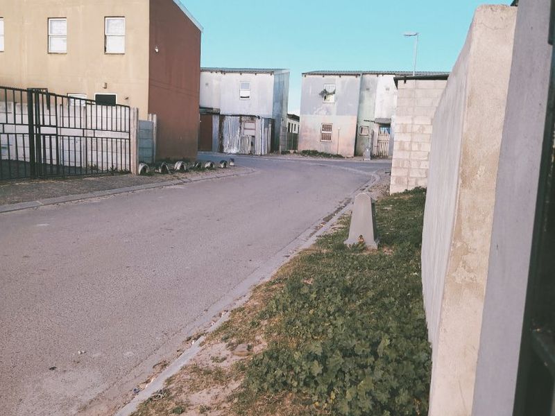 A street-level view of the residential townhouse complex, showing the access road, boundary walls, and surrounding building facades.