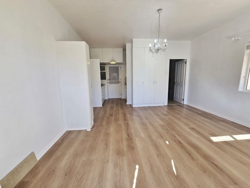 Spacious open-plan living area featuring light wood-look laminate flooring, built-in storage cupboards, and a decorative chandelier, leading into a functional kitchen space.