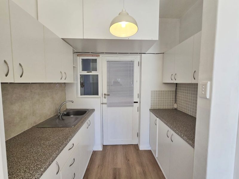 A functional galley-style kitchen featuring white cabinetry, laminate countertops, tiled splashbacks, and a stainless steel sink with direct access to an exterior door.