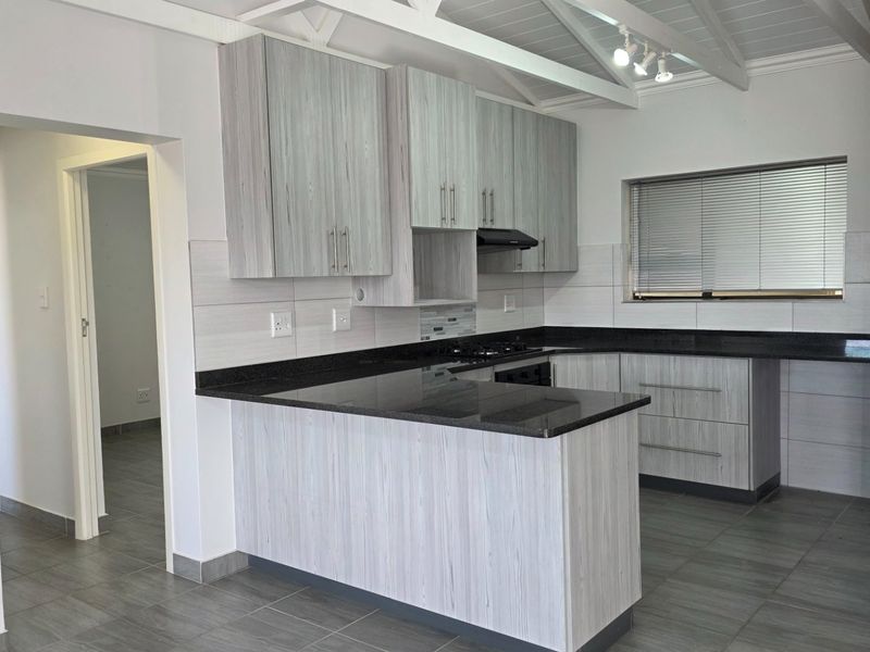 Modern kitchen featuring light wood-grain cabinetry, dark granite countertops, tiled flooring, and an exposed beam ceiling with track lighting.