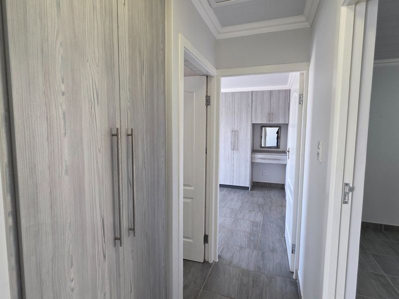 A clean hallway featuring modern wood-look built-in cupboards and neutral tiled flooring, leading into an adjacent bedroom area.