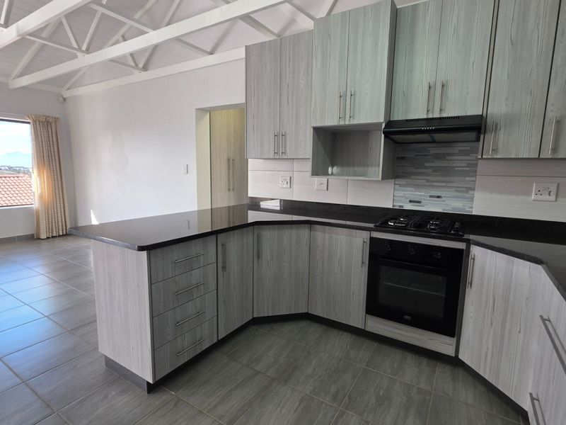 Modern open-plan kitchen featuring light wood-grain cabinetry, dark granite countertops, built-in oven, gas hob, and tiled flooring under exposed white roof trusses.