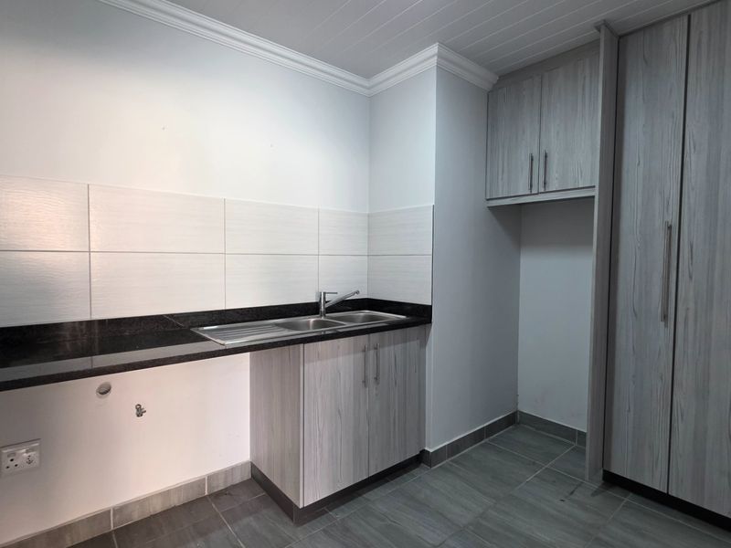 A clean, modern kitchen area featuring grey wood-grain cabinetry, granite countertops, a stainless steel sink, and neutral tiled finishes.