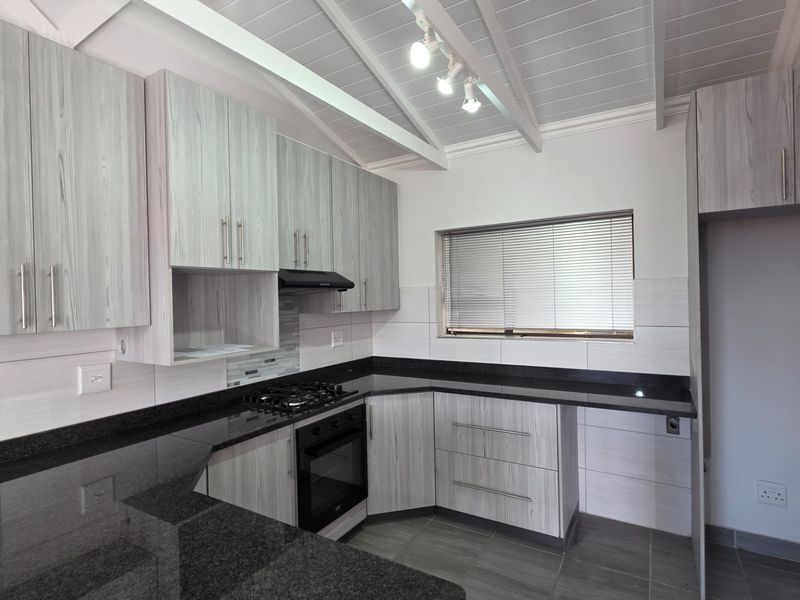 Modern kitchen featuring light wood-grain cabinetry, dark granite countertops, built-in oven with gas hob, and an airy exposed beam ceiling.