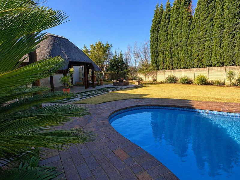 Spacious backyard featuring a swimming pool, brick-paved surround, and a traditional thatched lapa, set within a well-maintained garden with high boundary walls.