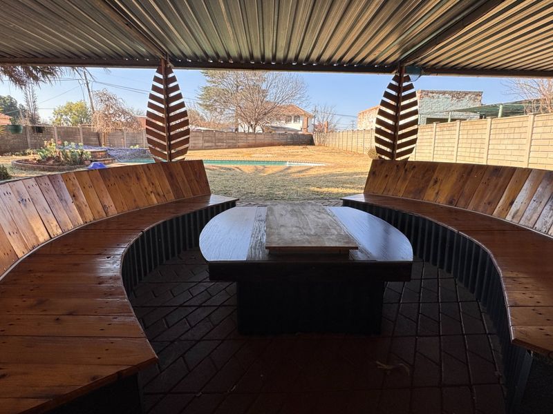 Covered outdoor patio featuring built-in curved wooden bench seating, corrugated metal roofing, and brick paving, overlooking the backyard and swimming pool area.