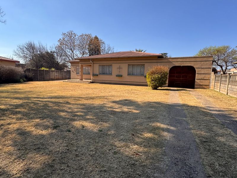 Front view of a single-story brick house featuring an attached garage, concrete driveway, and a large front lawn with perimeter boundary walling.