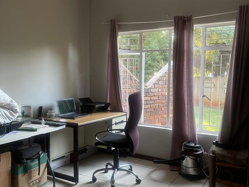 A functional home office space featuring tiled flooring and large windows with security bars, providing natural light and garden views.
