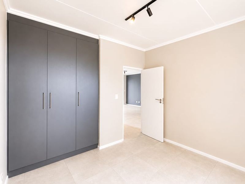 A bright, modern bedroom featuring neutral walls, large grey built-in cupboards, tiled flooring, and contemporary track lighting.