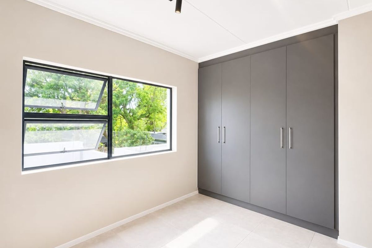 A bright, modern bedroom featuring neutral walls, large format floor tiles, and sleek floor-to-ceiling built-in cupboards with contemporary track lighting.