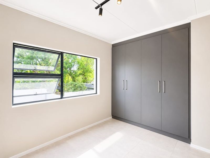 A bright, modern bedroom featuring neutral walls, large format floor tiles, and sleek floor-to-ceiling built-in cupboards with contemporary track lighting.
