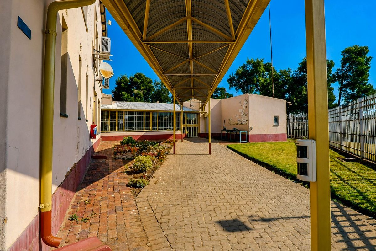 A paved rear exterior area featuring a covered walkway, security fencing, and access to the building structure with visible exterior wall maintenance required.