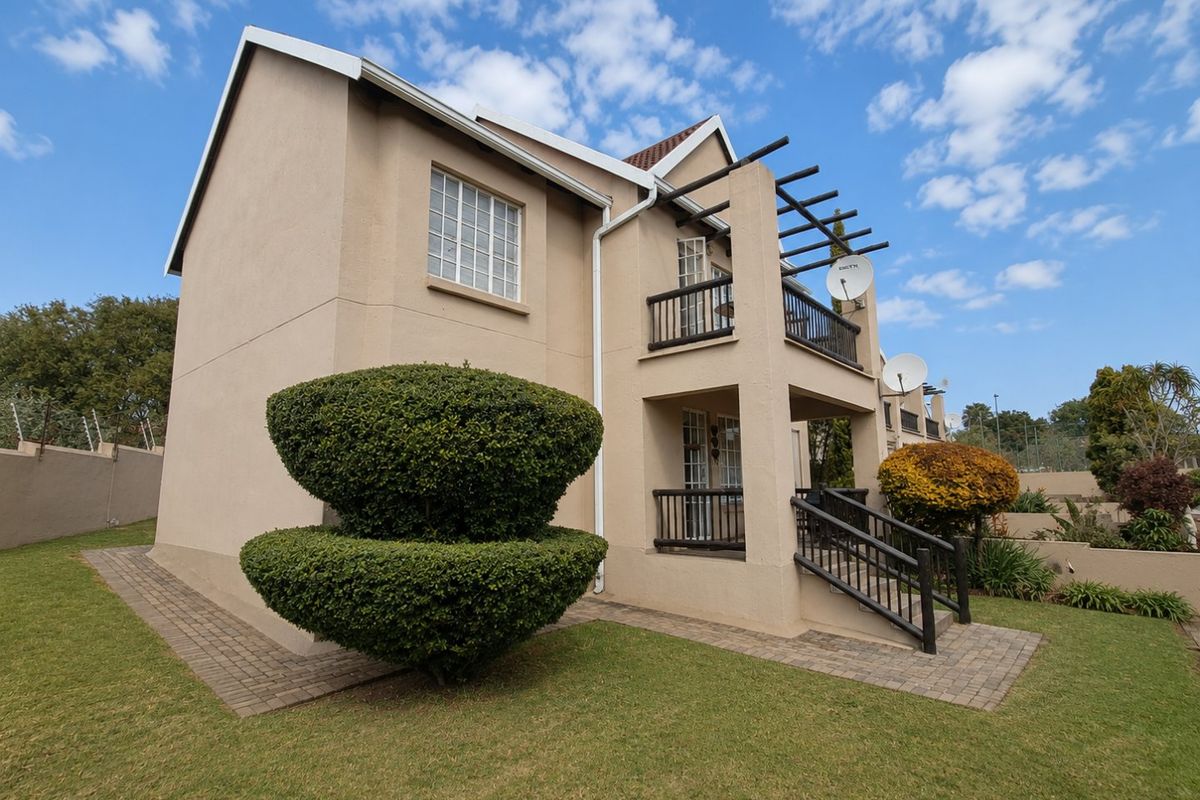 Rear view of the apartment building featuring a ground floor patio, upper level balcony with pergola, and paved access paths set within a landscaped garden.