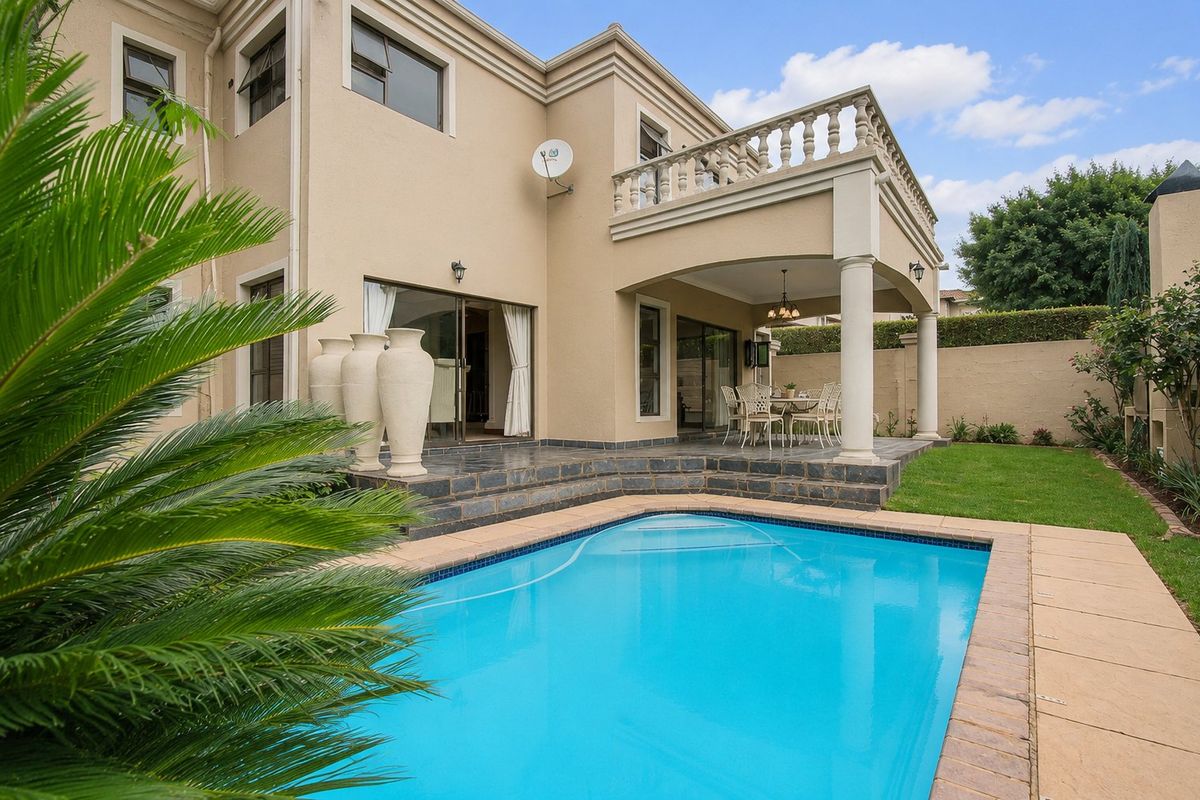 Rear exterior view featuring a swimming pool, covered patio area, and an upper-level balcony with stone paving and classic architectural detailing.