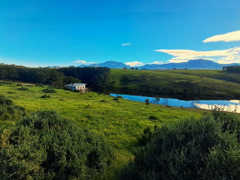 Scenic landscape view featuring a farm dam, rolling green pastures, and distant mountain ranges under a clear blue sky.