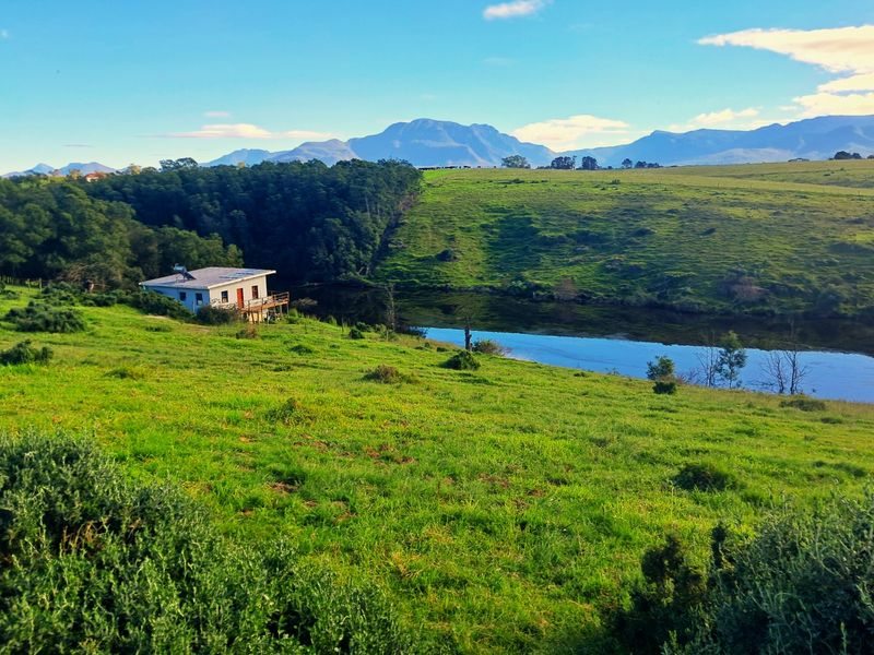 A scenic landscape view of a farm property featuring a dwelling with a wooden deck and solar water heater overlooking a tranquil dam.