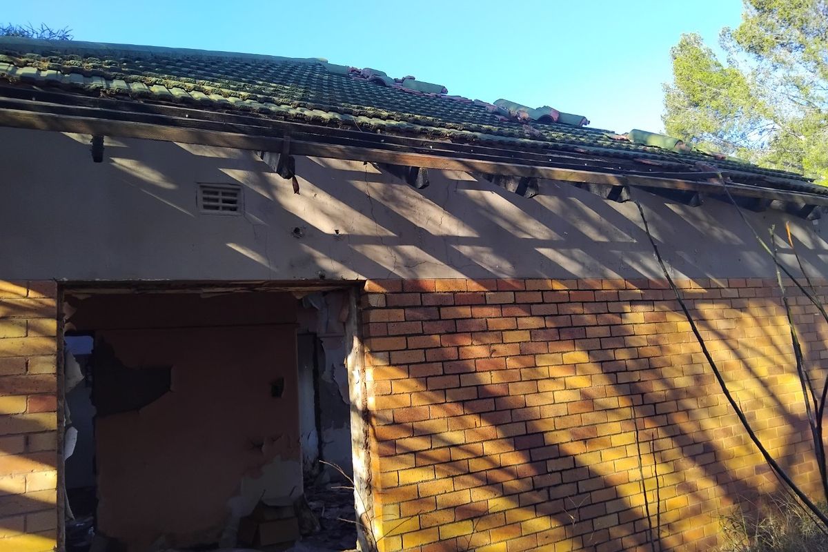 Exterior view of a dilapidated residential structure showing significant roof damage, missing tiles, exposed rafters, and damaged brickwork.