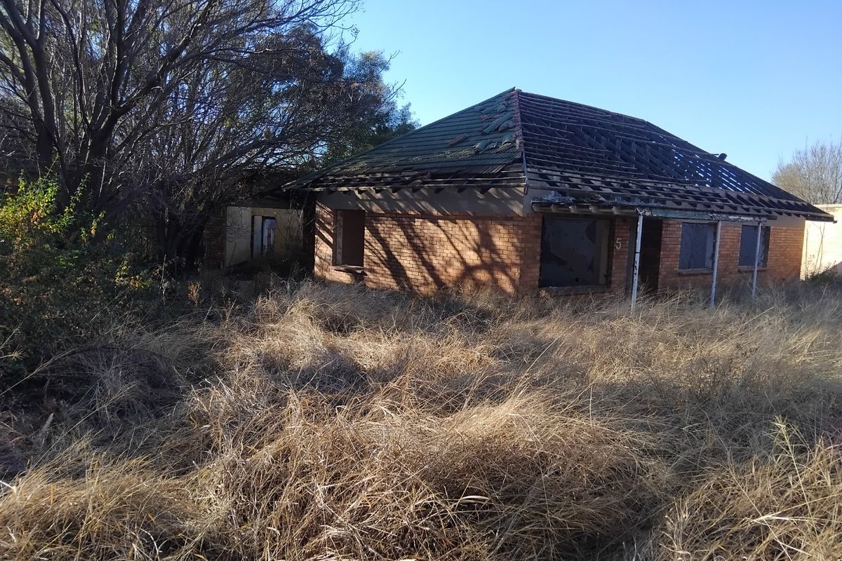 A dilapidated residential property featuring a brick facade and damaged roof structure, situated on an overgrown plot of land.