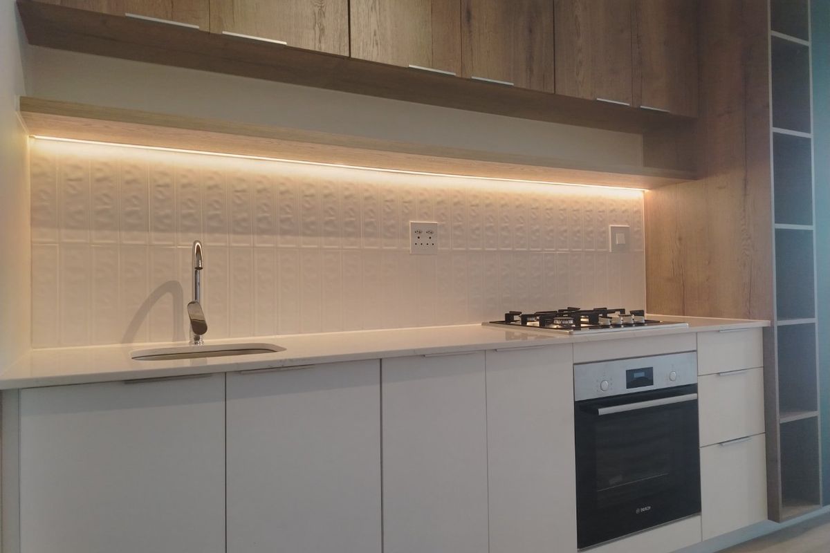 Modern kitchen featuring white cabinetry, stone countertops, integrated gas hob, built-in oven, and stylish under-cabinet lighting with a textured tiled backsplash.