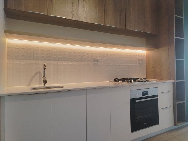 Modern kitchen featuring white cabinetry, stone countertops, integrated gas hob, built-in oven, and stylish under-cabinet lighting with a textured tiled backsplash.