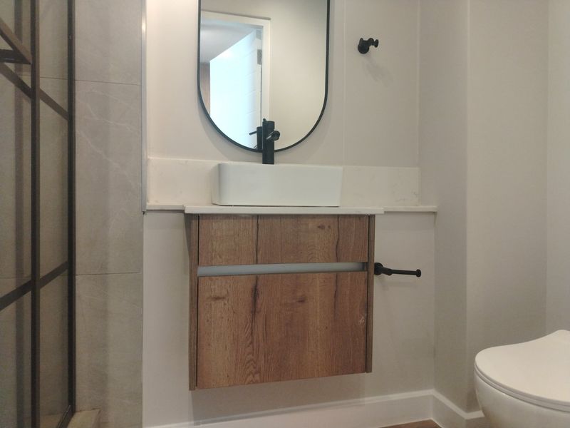 Modern bathroom featuring a wall-mounted wood-finish vanity, vessel sink with black fixtures, stone countertop, and a sleek black-framed mirror.