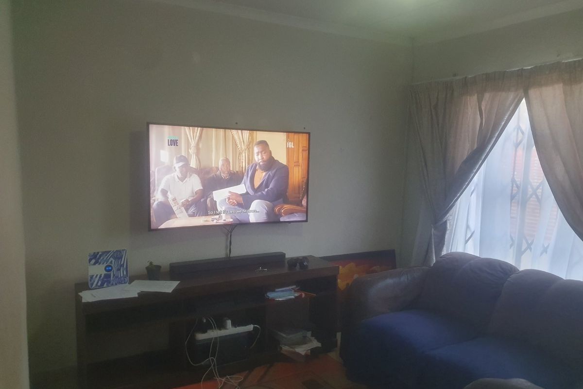 A living room featuring a wall-mounted television, curtained window with security gate, and neutral wall finishes.