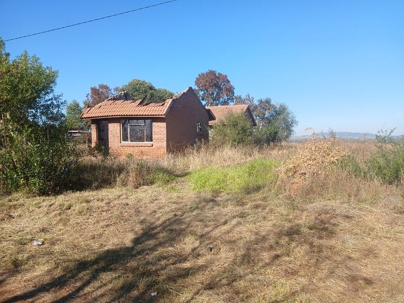 A view of a dilapidated brick structure on a vacant residential plot, showing significant roof damage and overgrown grounds.