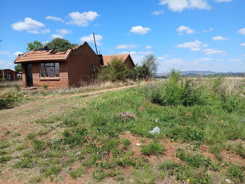 A view of a residential property showing a brick structure with significant roof damage and deterioration, situated on a large vacant plot.