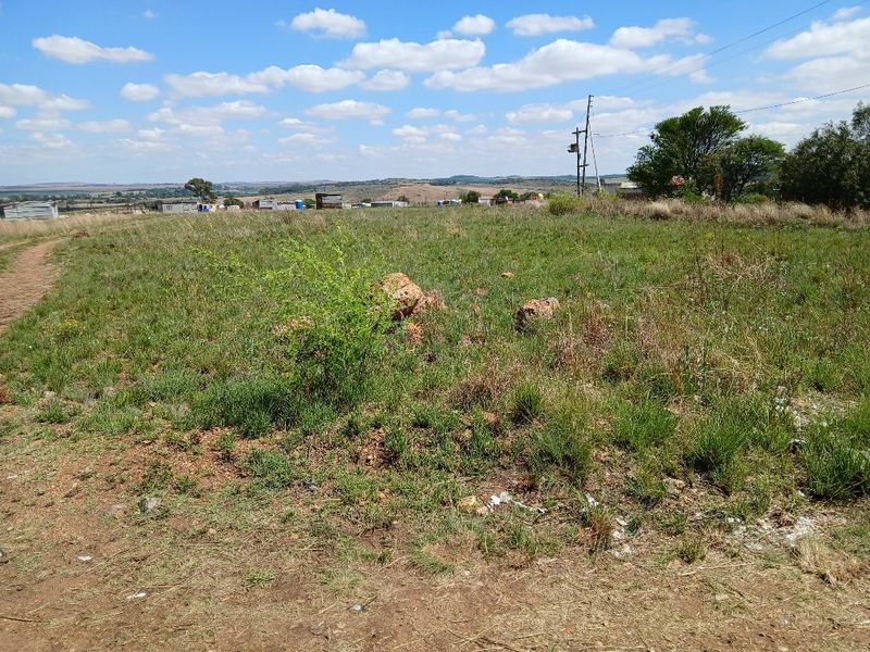 A view of a vacant residential plot featuring natural grass terrain, scattered rocks, and overhead utility infrastructure.