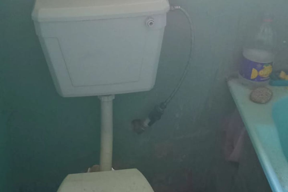 A small bathroom featuring a standard toilet and vanity basin with patterned floor tiles and painted walls requiring maintenance.