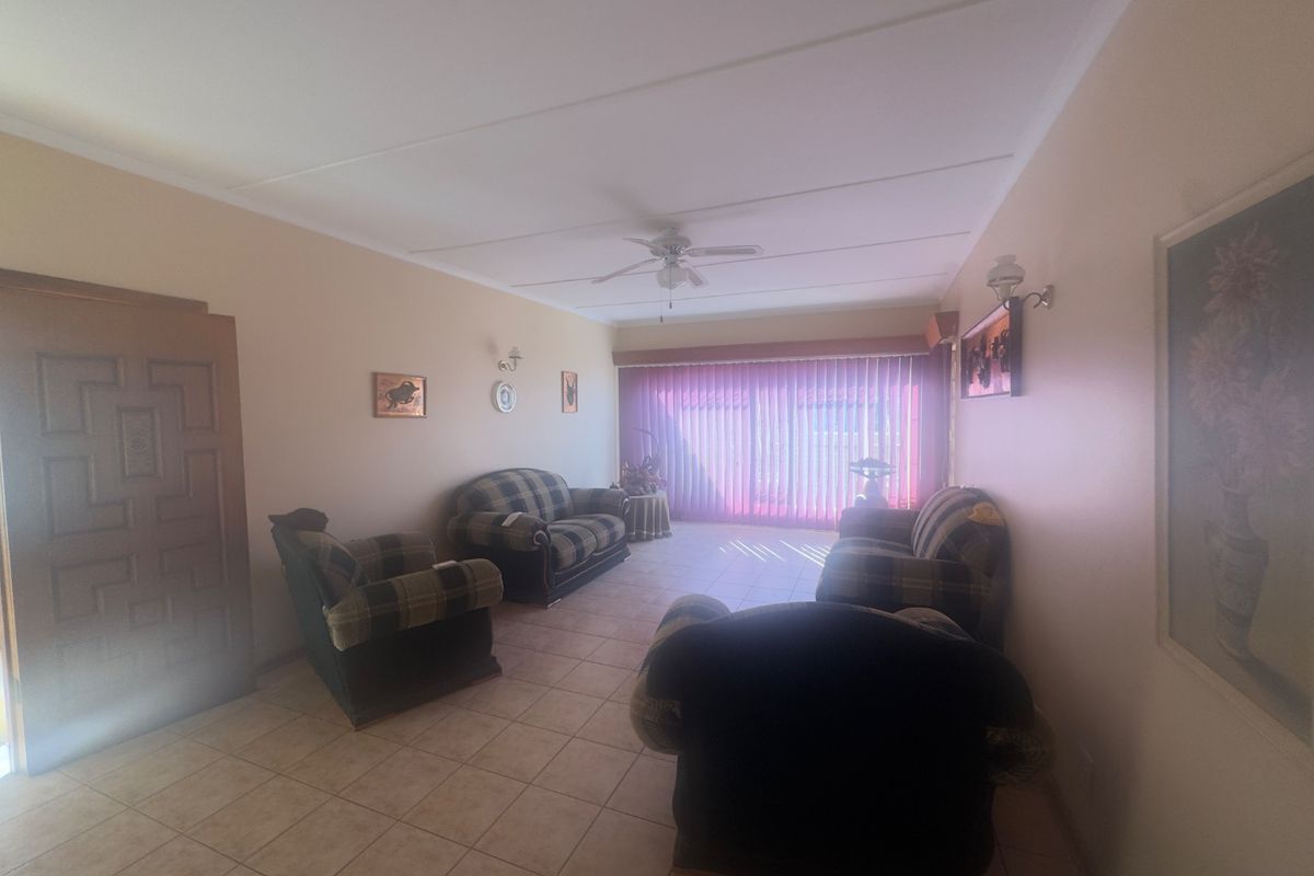 A living room featuring tiled flooring, a ceiling fan, and large windows covered by vertical blinds, providing a functional and neutral living space.