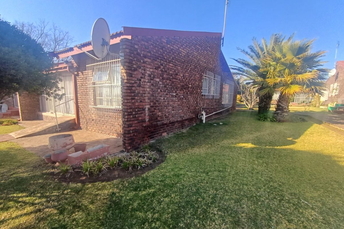 Rear view of a face brick townhouse featuring a paved patio area, security bars on windows, and a well-maintained garden with a palm tree.