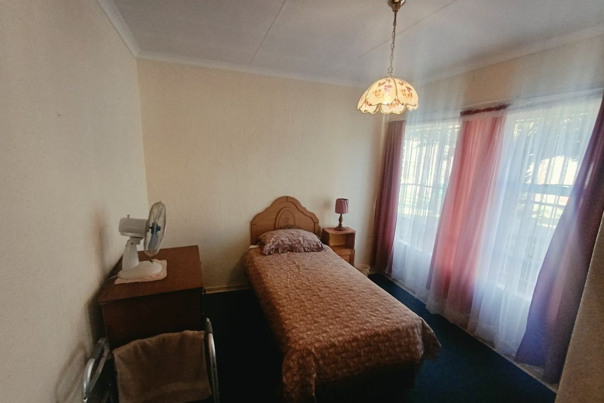 A functional bedroom featuring neutral walls, carpeted flooring, and a large window with curtain treatments, providing natural light.