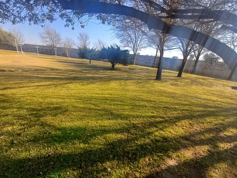 A spacious, open garden area featuring a well-maintained lawn, mature trees, and a secure perimeter wall.