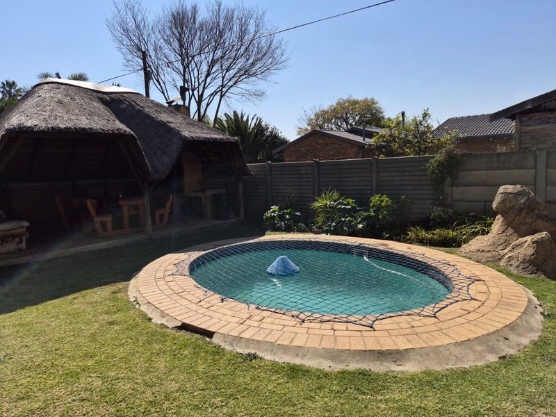 Outdoor entertainment area featuring a swimming pool with safety net, a thatched lapa with built-in braai, and a landscaped garden with boundary wall.