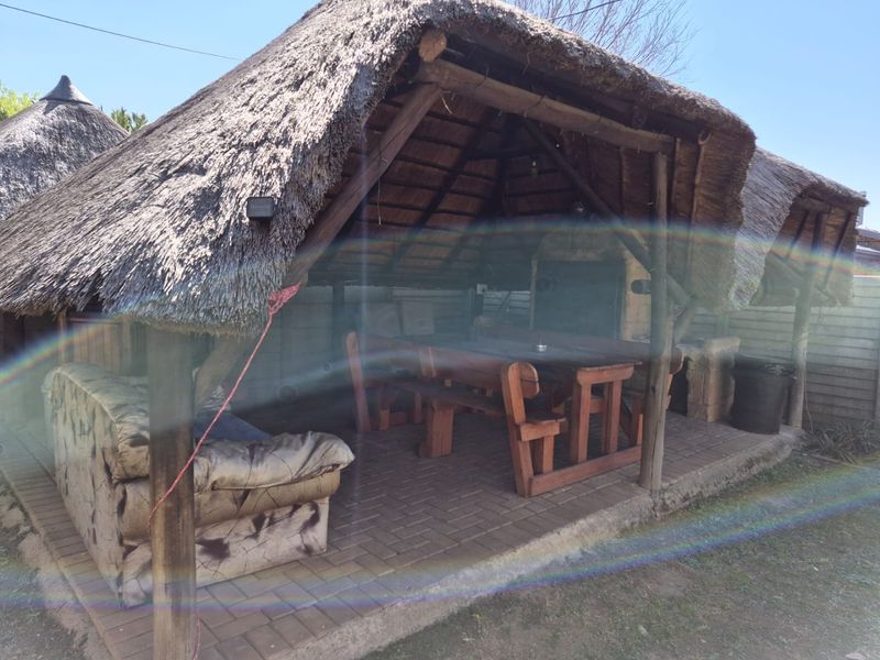 Outdoor entertainment area featuring a traditional thatch roof, timber support structure, brick paving, and a built-in braai facility.