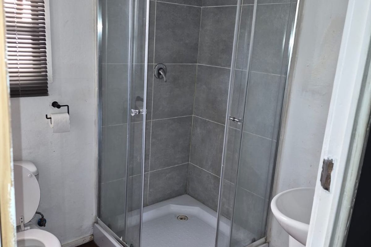 A compact bathroom featuring a glass-enclosed shower, pedestal basin, and toilet, with grey tiled shower walls and wooden flooring.