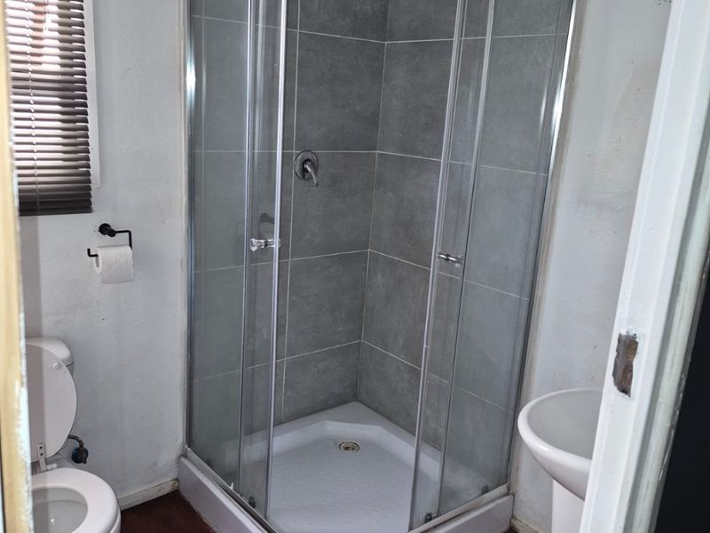 A compact bathroom featuring a glass-enclosed shower, pedestal basin, and toilet, with grey tiled shower walls and wooden flooring.