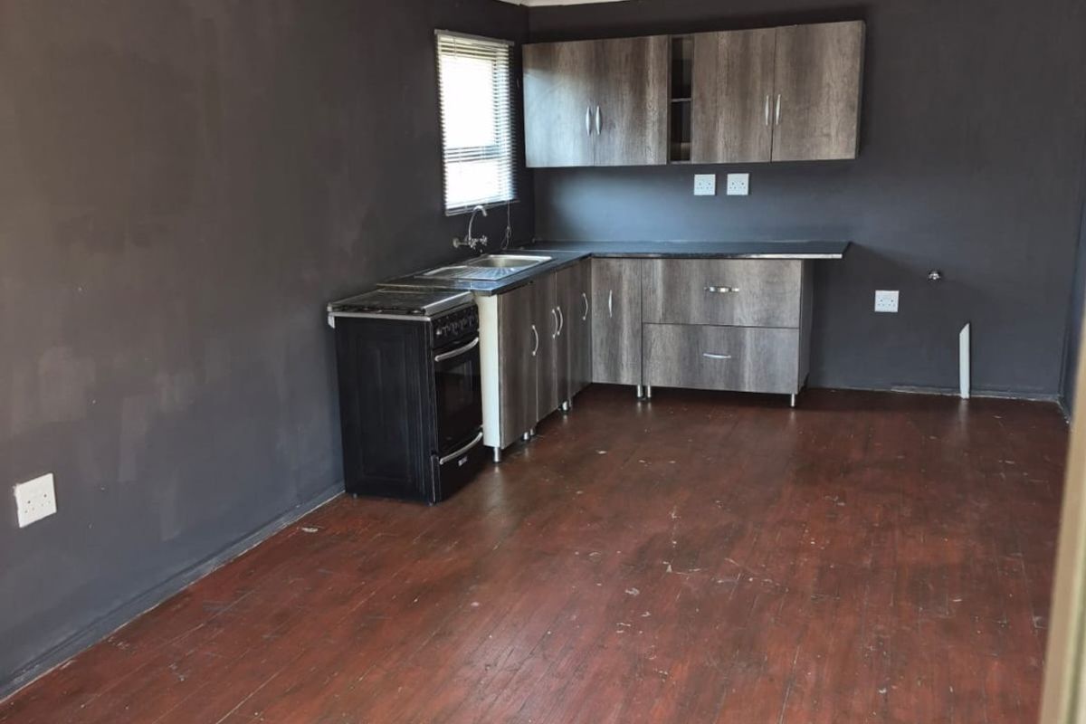 An empty kitchen featuring dark grey walls, wood-look cabinetry, a stainless steel sink, and worn timber flooring, offering a blank canvas for renovation.