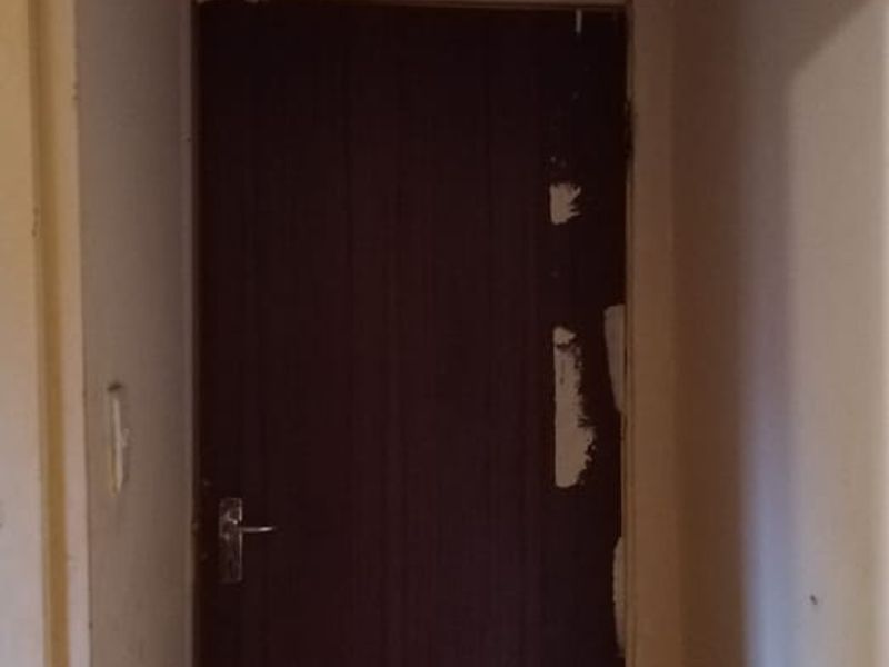 A narrow hallway featuring tiled flooring and a dark internal door, showing significant signs of wear including ceiling staining and damaged door finish.