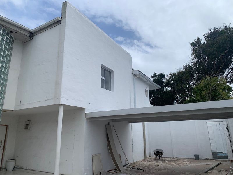 Rear exterior view of the property featuring a white rendered facade, glass block detailing, and a carport structure with ongoing exterior maintenance.