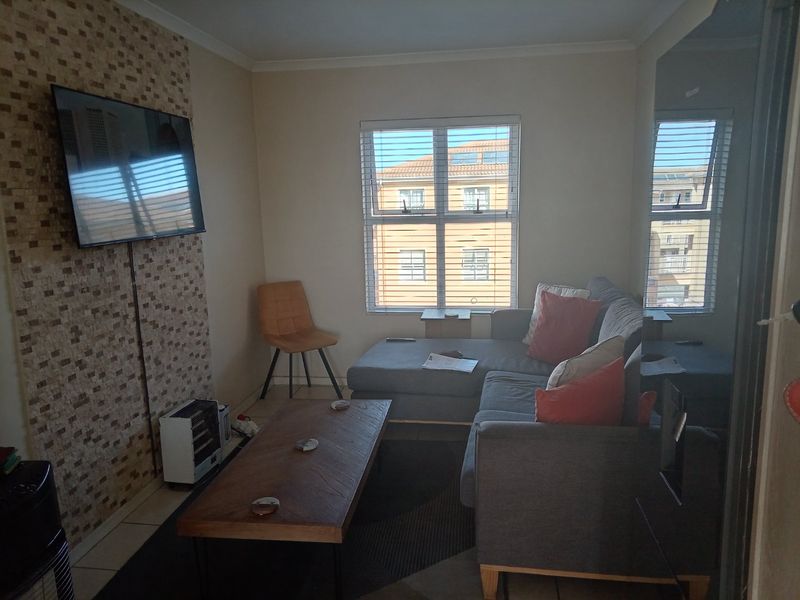 A comfortable living room featuring a tiled accent wall, tiled flooring, and large windows with venetian blinds allowing for natural light.