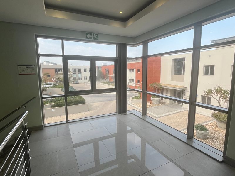 A bright, modern commercial hallway featuring expansive floor-to-ceiling windows, polished tile flooring, and a sleek stainless steel handrail overlooking the office complex courtyard.