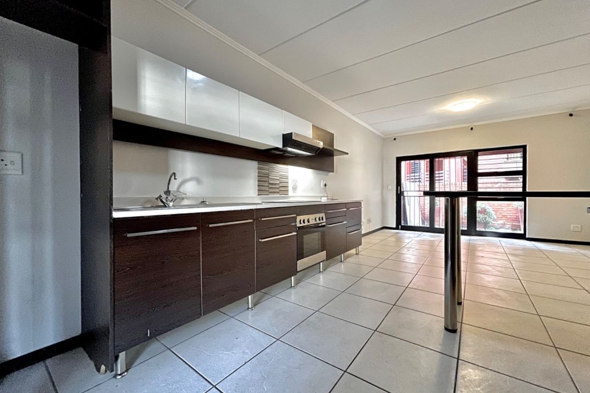 A modern, open-plan kitchen featuring dark wood cabinetry, white upper cabinets, stainless steel appliances, and tiled flooring, leading to an outdoor area.