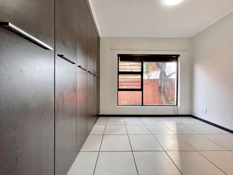 A bright, empty bedroom featuring large dark-toned built-in cupboards, neutral tiled flooring, and a window fitted with security bars.