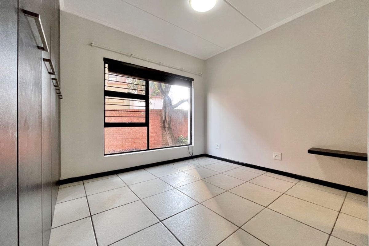 A clean, empty bedroom featuring tiled flooring, built-in cupboards, and a window with security bars.