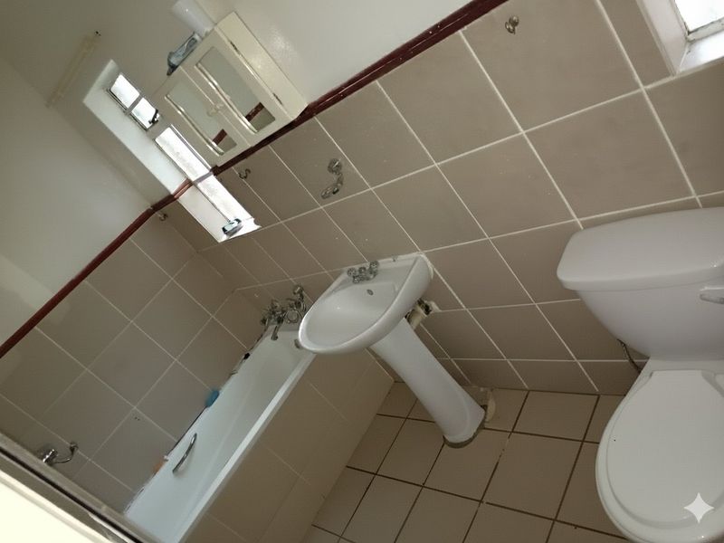 A dated bathroom featuring a pedestal basin, bathtub, and toilet with neutral wall and floor tiling, requiring cosmetic updates.